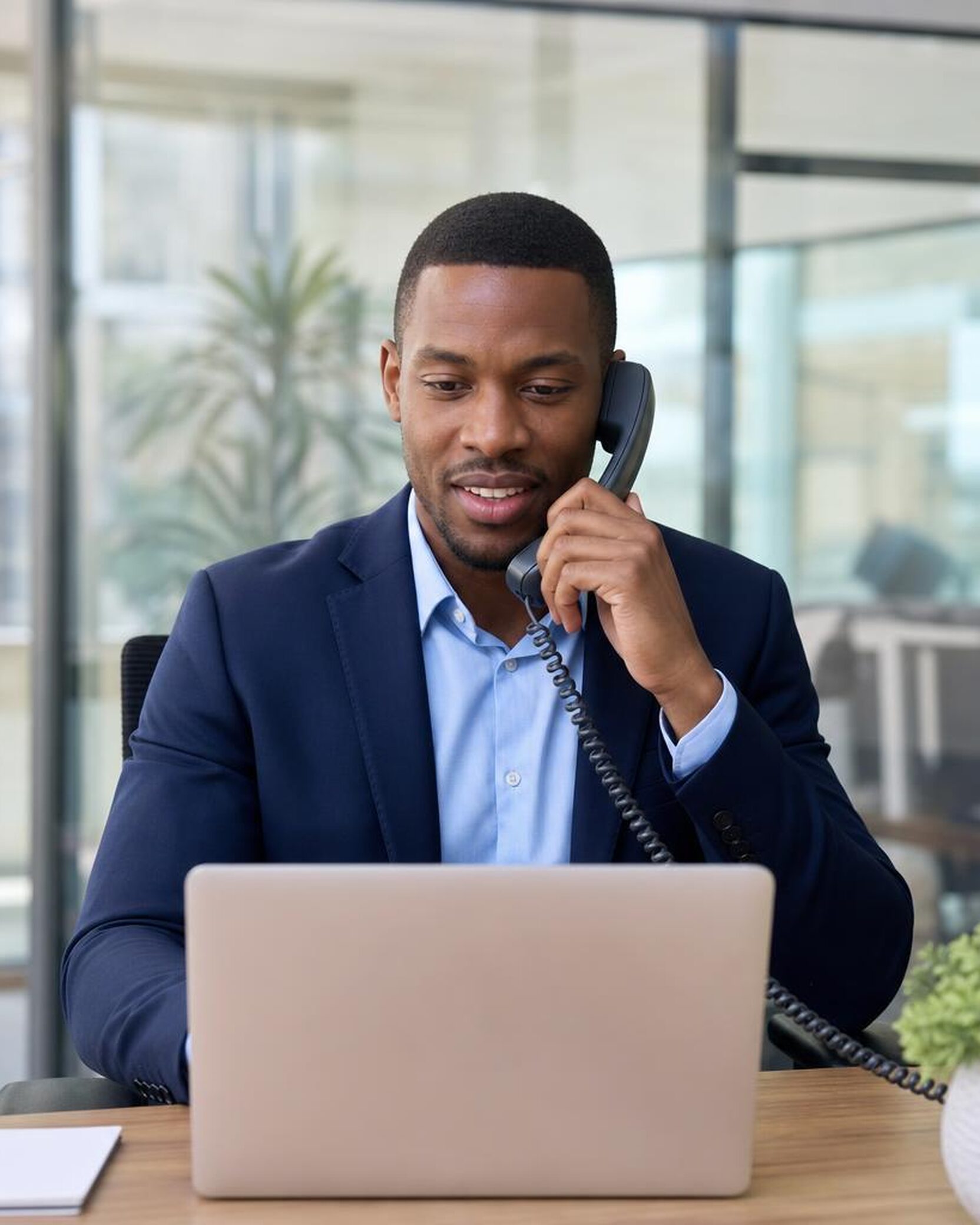 A relationship banker in his 30s in a navy suit and light blue shirt sits at a desk in a bright, modern bank office, phone to his ear.
