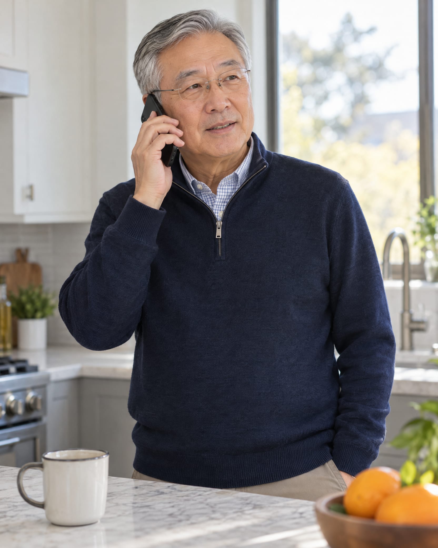 A Korean American man in his late 60s in a quarter-zip sweater stands in a sunlit kitchen, phone to his ear.